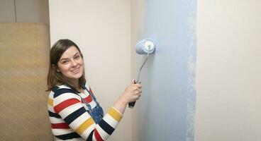 femme permanent habillé dans salopette et rayé chemisier, profil voir, souriant à caméra avec une rouleau dans sa main La peinture une blanc mur avec bleu peindre photo