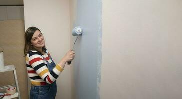 femme permanent habillé dans salopette et rayé chemisier, profil voir, souriant à caméra avec une rouleau dans sa main La peinture une blanc mur avec bleu peindre photo