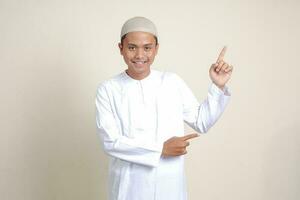 portrait de attrayant asiatique musulman homme dans blanc chemise avec calotte montrant produit et montrer du doigt avec le sien main et doigt à le côté. La publicité concept. isolé image sur gris Contexte photo