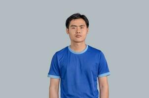 Jeune asiatique homme dans bleu t-shirts permanent dans studio et sérieux concentré visage à caméra, à la recherche agressif, isolé sur gris Contexte. détendu avec sérieux expression sur affronter. photo