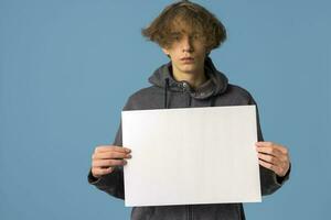 une sérieux, mécontent adolescent dans une gris sweat à capuche et ondulé cheveux détient un vide affiche sur une bleu Contexte. photo