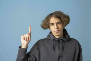 une adolescent dans une gris sweat à capuche avec ondulé cheveux points en haut avec le sien indice doigt sur une bleu Contexte photo