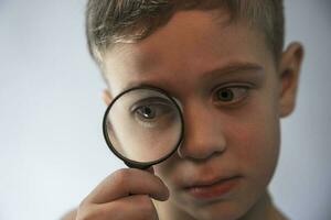 une mignonne caucasien garçon avec une grossissant verre à le sien œil est en jouant détective. photo