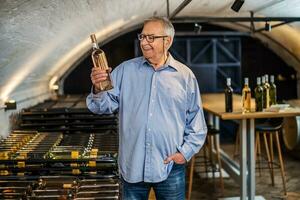 portrait de Sénior homme qui possède vignoble. il est examiner qualité dans le sien du vin cave. industrie du vin fabrication concept. photo