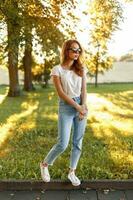 élégant jolie fille dans taille haute jeans, blanc T-shirt et baskets dans une parc sur une Contexte de des arbres à le coucher du soleil. photo