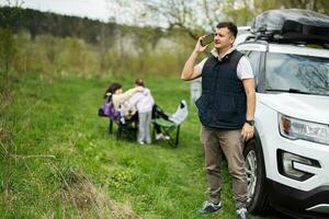 homme dans Sans manches gilet parler sur téléphone contre suv voiture avec toit grille boîte, famille sur pique-nique. photo