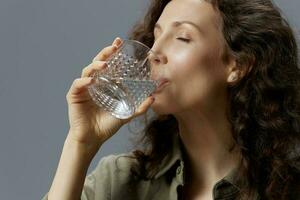 fermer portrait de frisé magnifique femme dans décontractée kaki vert chemise tenir verre prendre plaisir nettoyer l'eau posant isolé sur plus de gris bleu Contexte. en bonne santé mode de vie. l'eau est la vie concept. copie espace photo