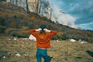 une femme dans une chandail détient sa mains derrière sa tête et regards à le haute montagnes paysage bleu ciel des nuages photo