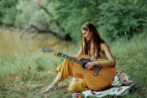 hippie femme en jouant guitare souriant et en chantant Chansons dans la nature séance sur une plaid par le Lac dans le soir dans le des rayons de le réglage Soleil. une mode de vie dans harmonie avec le corps et la nature photo