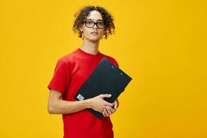 confus myope Jeune étudiant homme dans rouge T-shirt marrant lunettes détient tablette dossier avec étude Remarques posant isolé sur plus de Jaune studio Contexte. gratuit endroit pour publicité. éducation Université concept photo