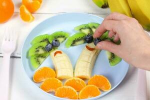 Créatif nourriture pour bébé, kiwi, mandarines et banane, marrant nourriture sur coloré vaisselle. fruit pour une enfant. Haut voir, plat allonger photo