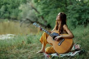 Jeune hippie femme séance en plein air sur une plaid et en jouant sa guitare sur le berge de rivière dans le le coucher du soleil photo