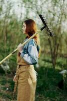 femme magnifiquement souriant agriculteur dans travail vêtements et tablier travail en plein air dans la nature et en portant une râteau à recueillir herbe photo