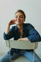 une Jeune femme séance dans une chaise à Accueil souriant avec les dents avec une court la Coupe de cheveux dans jeans et une denim chemise sur une blanc Contexte. fille Naturel pose avec non filtres photo