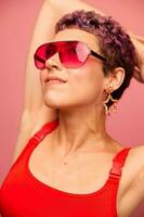 mode portrait de une femme avec une court la Coupe de cheveux dans coloré des lunettes de soleil avec inhabituel accessoires avec des boucles d'oreilles souriant sur une rose brillant Contexte photo