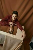 une homme avec une miroir est assis à une table sur une en tissu Contexte et une marron chemise photo