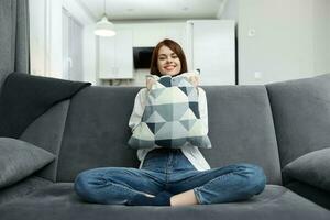 souriant femme avec une oreiller dans sa mains est assis sur canapés dans un appartement photo