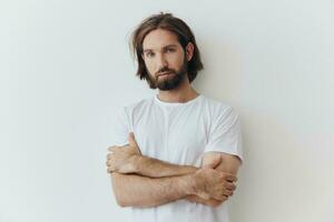 une homme avec une barbe et longue cheveux dans une blanc T-shirt et bleu jeans des stands contre une blanc mur penché toute la longueur sur il, détendu style photo