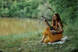 une hippie femme en jouant sa guitare sourit et chante Chansons dans la nature séance sur une plaid dans le soir dans le le coucher du soleil lumière du soleil. une mode de vie dans harmonie avec le corps et la nature photo