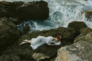 magnifique femme dans longue blanc robe humide cheveux mensonge sur une rocheux falaise la nature paysage photo