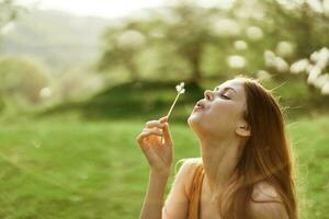 le content femme sourit et coups le pissenlit dans le vent. été vert paysage et ensoleillement dans le Contexte photo