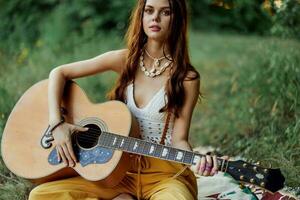 hippie femme souriant et étreindre sa guitare dans la nature dans le parc dans le le coucher du soleil lumière dans éco-vêtements photo