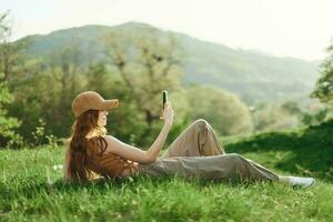 Haut vue une Jeune femelle free-lance étudiant dans une Jaune casquette, haut, et vert un pantalon est travail sur sa cellule téléphone séance sur le vert herbe dans le parc. mode de vie et concept de une Jeune en bonne santé société photo