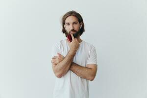 une homme avec une barbe et longue cheveux dans une blanc T-shirt et bleu jeans des stands contre une blanc mur, penché contre il et écoute à la musique avec sans fil blanc écouteurs, regarder pensivement photo