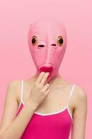 marrant femme dans une sexy costume avec une rose poisson masque sur sa tête mettre sa les doigts dans sa bouche dans rose vêtements sur une rose Contexte. le concept de moderne art la photographie photo