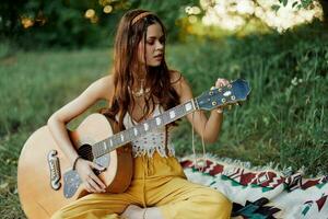 fille hippie femme en jouant guitare dans respectueux de la nature vêtements séance sur le sol à l'extérieur dans la nature dans le tomber en train de regarder le le coucher du soleil photo