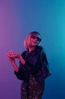 de bonne humeur sexy magnifique blond femme dans cuir veste scintillant robe branché des lunettes de soleil plier mains posant isolé dans bleu rose Couleur lumière studio Contexte. néon fête cyberpunk concept. copie espace photo