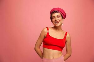 Jeune athlétique femme avec une court la Coupe de cheveux et violet cheveux dans une rouge Haut et une rose chapeau avec un athlétique figure sourit et grimaces à la recherche à le caméra sur une rose Contexte photo