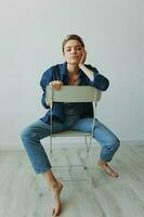 une Jeune femme séance dans une chaise à Accueil souriant avec les dents avec une court la Coupe de cheveux dans jeans et une denim chemise sur une blanc Contexte. fille Naturel pose avec non filtres photo