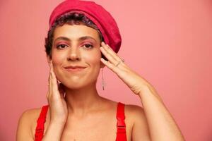 Jeune athlétique femme avec une court la Coupe de cheveux et violet cheveux dans une rouge Haut et une rose chapeau avec un athlétique figure sourit et grimaces montrant panneaux avec sa mains à la recherche à le caméra sur une rose Contexte photo