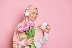 délicieux arabe musulman femme dans rose hijab, en train de lire salutation carte tandis que avoir une bouquet de tulipes pour de la mère journée photo
