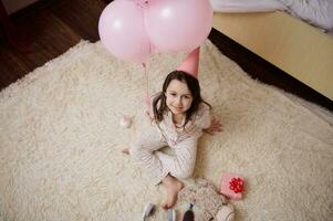 Haut vue peu enfant fille dans élégant pyjamas et rose de fête chapeau, séance sur tapis et souriant à la recherche en haut à caméra photo