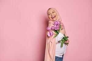 Enceinte arabe musulman femme dans hijab détient une bouquet de tulipes pour de la mère jour, sourit à la recherche de côté, rose Contexte photo