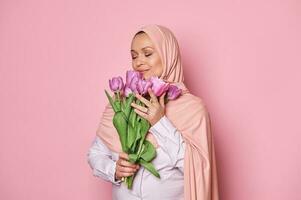 charmant musulman femme dans rose hijab, reniflement bouquet de tulipes, posant avec sa yeux fermé sur isolé rose Contexte photo