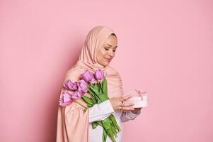 souriant musulman femme dans rose hijab, en portant une bouquet de tulipes et mignonne cadeau boîte pour spécial occasion, isolé sur rose photo