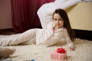 adorable peu fille avec longue cheveux, habillé dans élégant pyjamas avec multicolore des points, mensonge sur tapis dans sa chambre photo