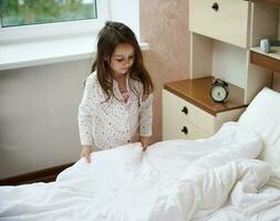 peu fille dans pyjama, pliant sa lit et nettoie le pièce après veille en haut dans le Matin. enseignement les enfants à commande photo