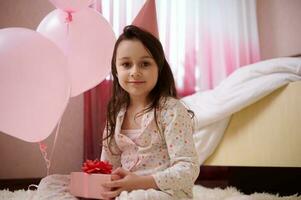 content peu enfant fille dans pyjamas et rose fête chapeau, détient une mignonne cadeau boîte pour anniversaire et sourit à la recherche à caméra photo
