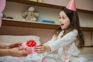 étonné anniversaire fille dans pyjamas, séance sur lit, exprimer content émotions tandis que recevoir mignonne présent pour sa anniversaire photo