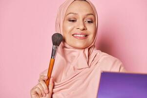 arabe musulman jolie femme dans rose hijab, sourit à la recherche à sa réflexion dans cosmétique miroir tandis que s'applique maquillage sur visage photo