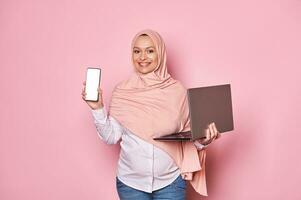 souriant Enceinte arabe musulman femme dans rose hijab, en portant téléphone intelligent avec blanc Vide filtrer, isolé rose Contexte photo