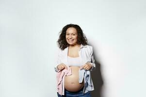 souriant Enceinte femme en portant bleu et rose nouveau née bodys près sa ventre. attendant pour une bébé, garçon ou fille, jumeaux photo