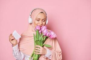 musulman femme dans rose hijab et écouteurs, en portant enveloppe avec salutation carte et reniflement une mignonne bouquet de tulipes photo