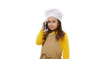 charmant enfant fille, peu boulanger confiseur dans du chef chapeau et beige tablier, parlant sur mobile téléphone, souriant à caméra photo