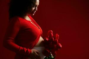 tondu vue de une Enceinte femme, femme enceinte mère émouvant sa ventre, posant avec rouge tulipes, isolé sur rouge toile de fond photo