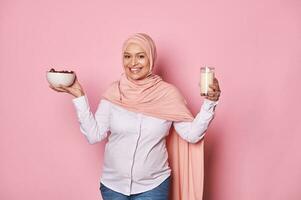 magnifique arabe musulman femme dans rose hijab, détient en bonne santé petit déjeuner dans Ramadan - bol avec sucré Rendez-vous et verre avec Lait photo